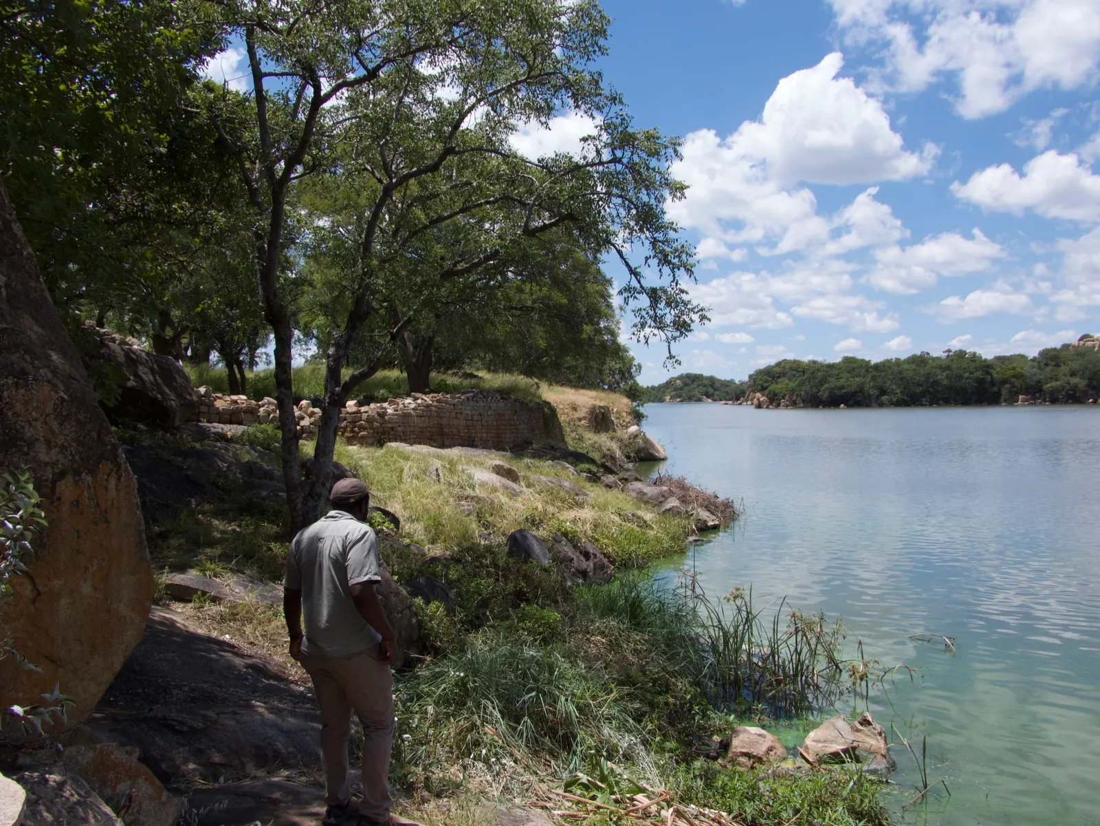 A scout standing at the edge of a dam at Khami, granite and trees across the water
